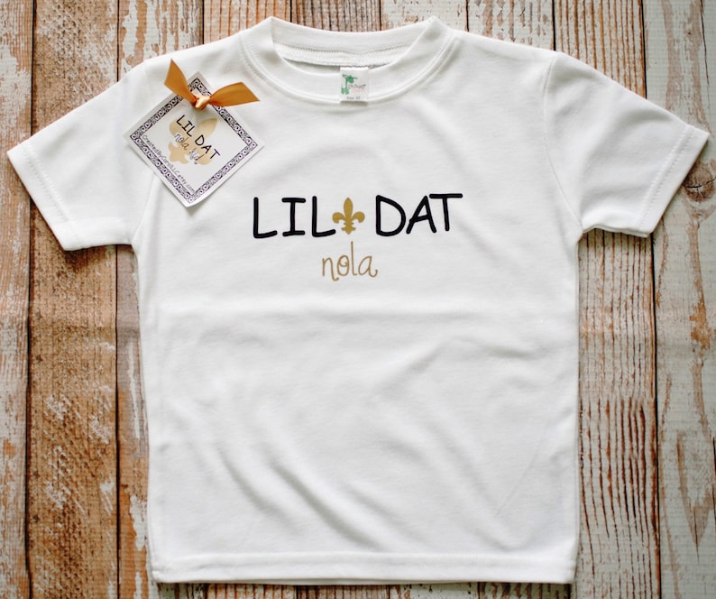 May include: White t-shirt with black text that reads "Lil Dat" with a gold fleur-de-lis and "nola" in gold lettering.