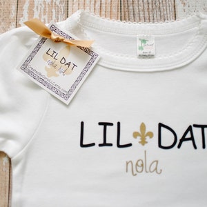 May include: White baby onesie with black text that reads "Lil Dat Nola" and a gold fleur-de-lis symbol.