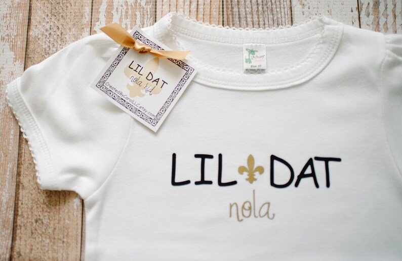 May include: White baby onesie with black text that reads "Lil Dat Nola" and a gold fleur-de-lis symbol.