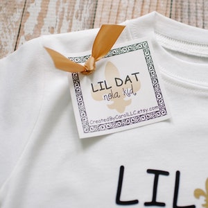 May include: White t-shirt with black text that reads "Lil Dat Nola Kid" and a gold fleur-de-lis. A white tag with black text and a gold ribbon reads "Lil Dat Nola Kid" and "CreatedByCarolLLC.etsy.com".