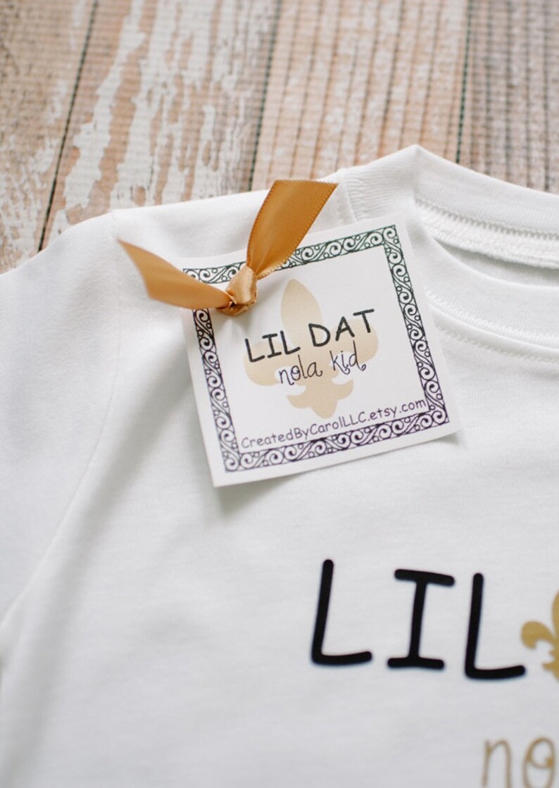 May include: White t-shirt with black text that reads "Lil Dat Nola Kid" and a gold fleur-de-lis. A white tag with black text and a gold ribbon reads "Lil Dat Nola Kid" and "CreatedByCarolLLC.etsy.com".