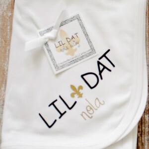 May include: White baby blanket with black embroidery that says "Lil Dat" and a gold fleur-de-lis. The blanket also has gold embroidery that says "nola".