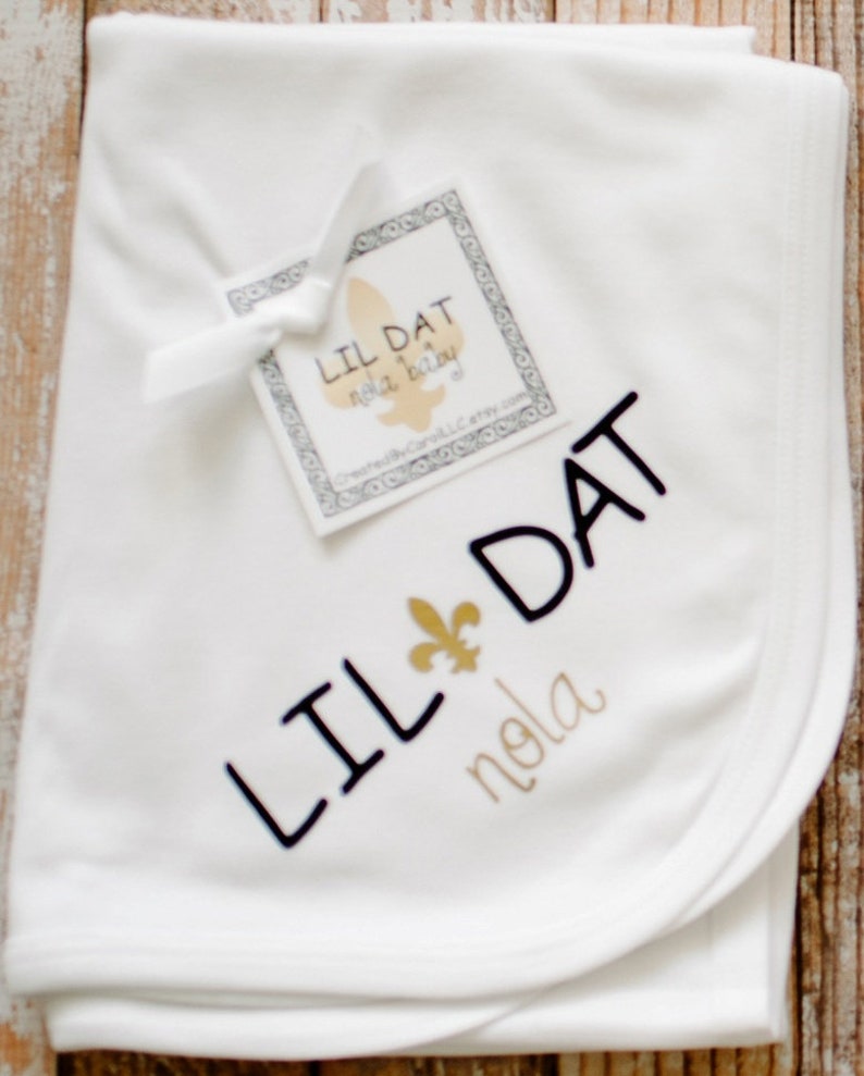 May include: White baby blanket with black embroidery that says "Lil Dat" and a gold fleur-de-lis. The blanket also has gold embroidery that says "nola".