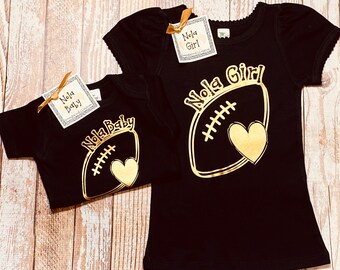May include: Two black t-shirts with gold foil designs. The larger shirt says "Nola Girl" with a football design. The smaller shirt says "Nola Baby" with a football design.