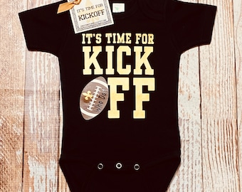 May include: Black bodysuit with gold text that reads "It's time for Kick Off" and a gold football with a fleur-de-lis on it. The text "Who Dat" is on the football.