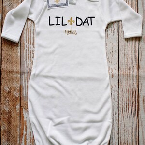 May include: White long-sleeved baby gown with gold lettering that reads "Lil Dat Nola" and a gold fleur-de-lis symbol.