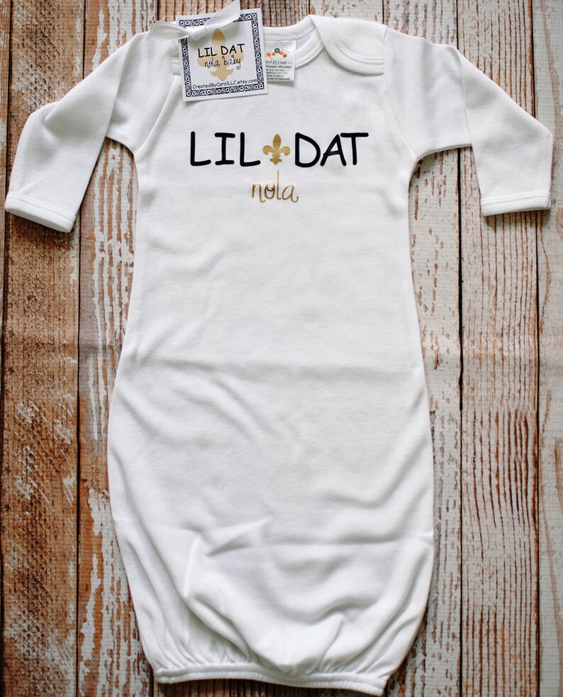 May include: White long-sleeved baby gown with gold lettering that reads "Lil Dat Nola" and a gold fleur-de-lis symbol.