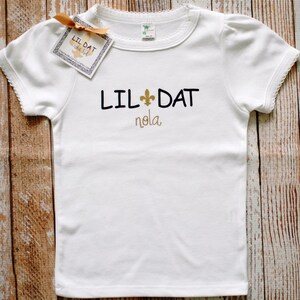 May include: White t-shirt with a black and gold design that says "Lil Dat Nola" with a fleur-de-lis symbol.
