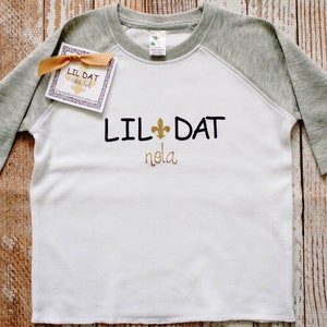 May include: White and grey baseball tee shirt with the text "Lil Dat" and a fleur-de-lis symbol in gold. The text "nola" is written below the main text.