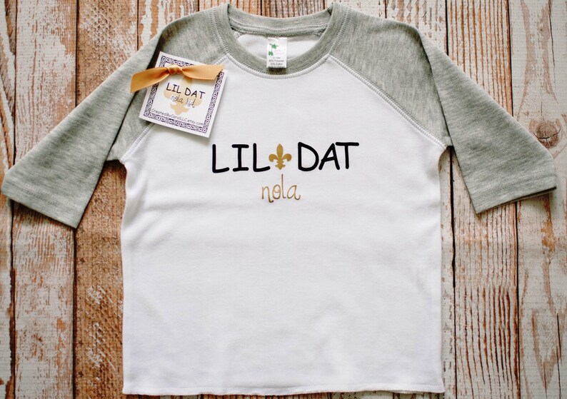 May include: White and grey baseball tee shirt with the text "Lil Dat" and a fleur-de-lis symbol in gold. The text "nola" is written below the main text.