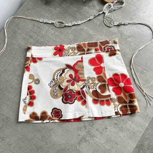 May include: A white mini skirt with a floral pattern in red, brown, and tan. The skirt has a pleated front and a matching silver belt with a decorative buckle. The belt is made of a thin, woven material.