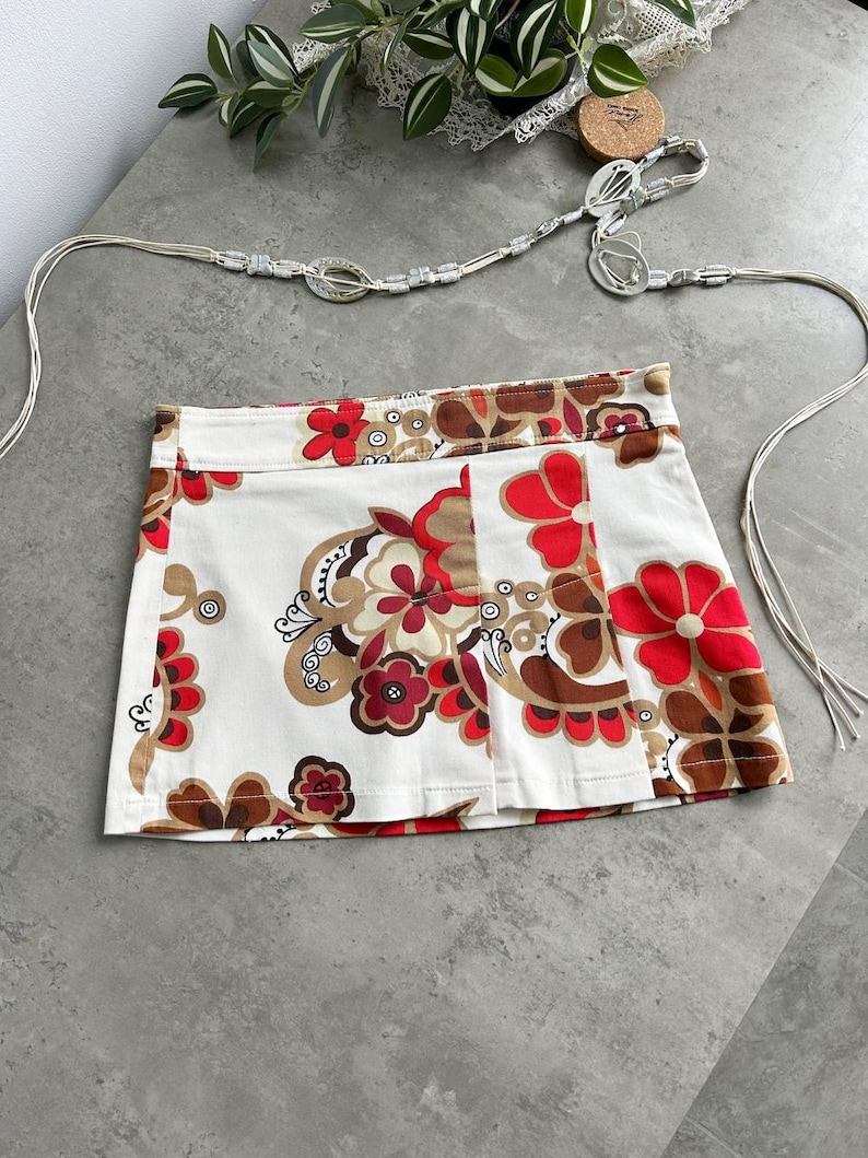 May include: A white mini skirt with a floral pattern in red, brown, and tan. The skirt has a pleated front and a matching silver belt with a decorative buckle. The belt is made of a thin, woven material.