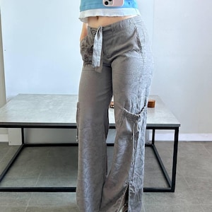 May include: Grey cargo trousers with a floral pattern, side pockets and a tie belt. The trousers have a wide leg design and a slit at the ankle. The model is wearing white shoes and a blue top.