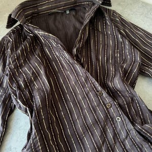 May include: A dark brown button-down shirt with vertical gold stripes. The long-sleeved shirt has a collared neckline and three visible buttons. The fabric appears to be a lightweight material.