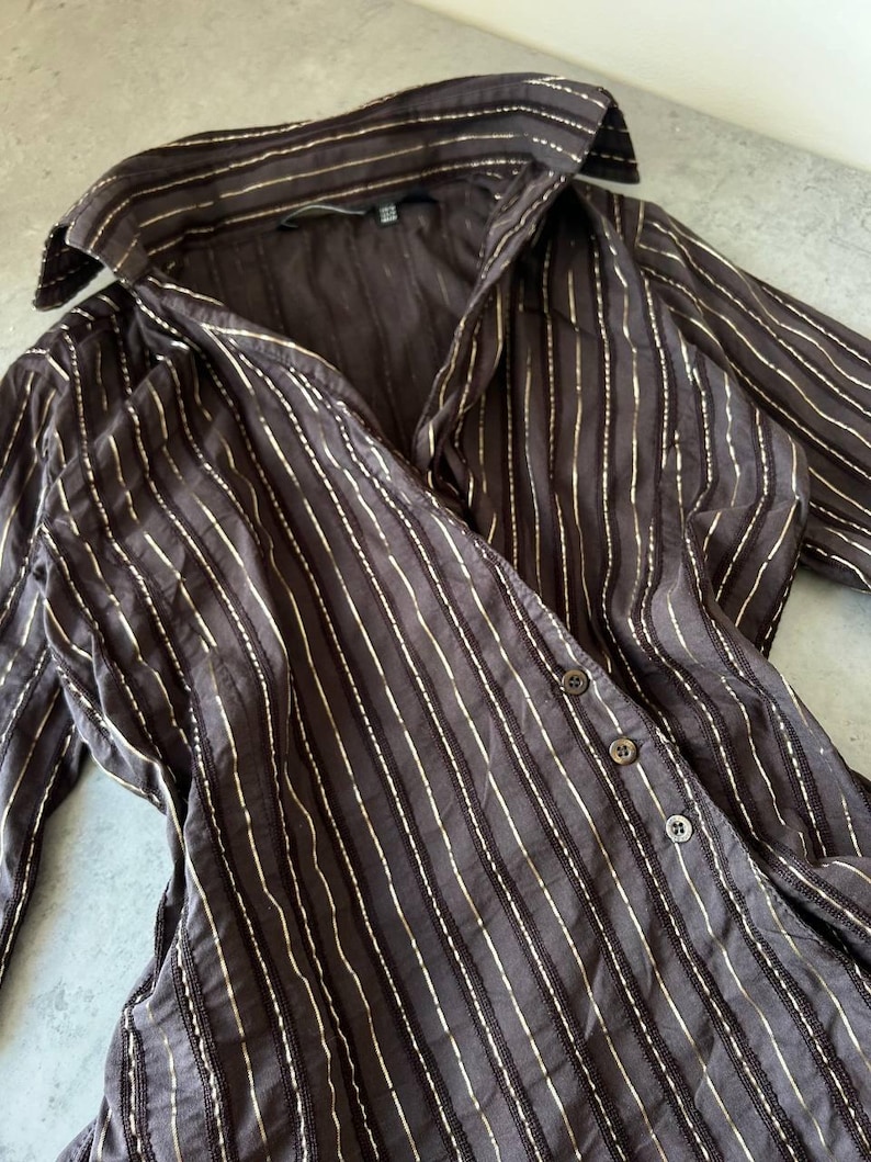 May include: A dark brown button-down shirt with vertical gold stripes. The long-sleeved shirt has a collared neckline and three visible buttons. The fabric appears to be a lightweight material.