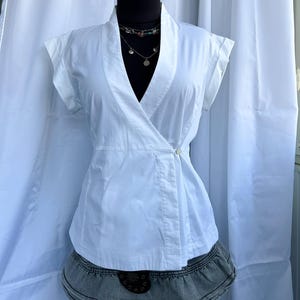 Peut inclure: Un haut blanc de style cache-cœur avec des manches courtes et une fermeture boutonnée. Le haut est associé à une jupe en jean. Un collier avec plusieurs rangs et pendentifs est visible.