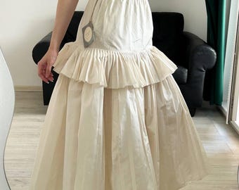 May include: A cream-colored maxi skirt featuring a high-waisted design with a button closure, a wide horizontal pleated ruffle detail around the mid-section, and a long, flowing, pleated skirt that extends to the ankles, paired with a light top.