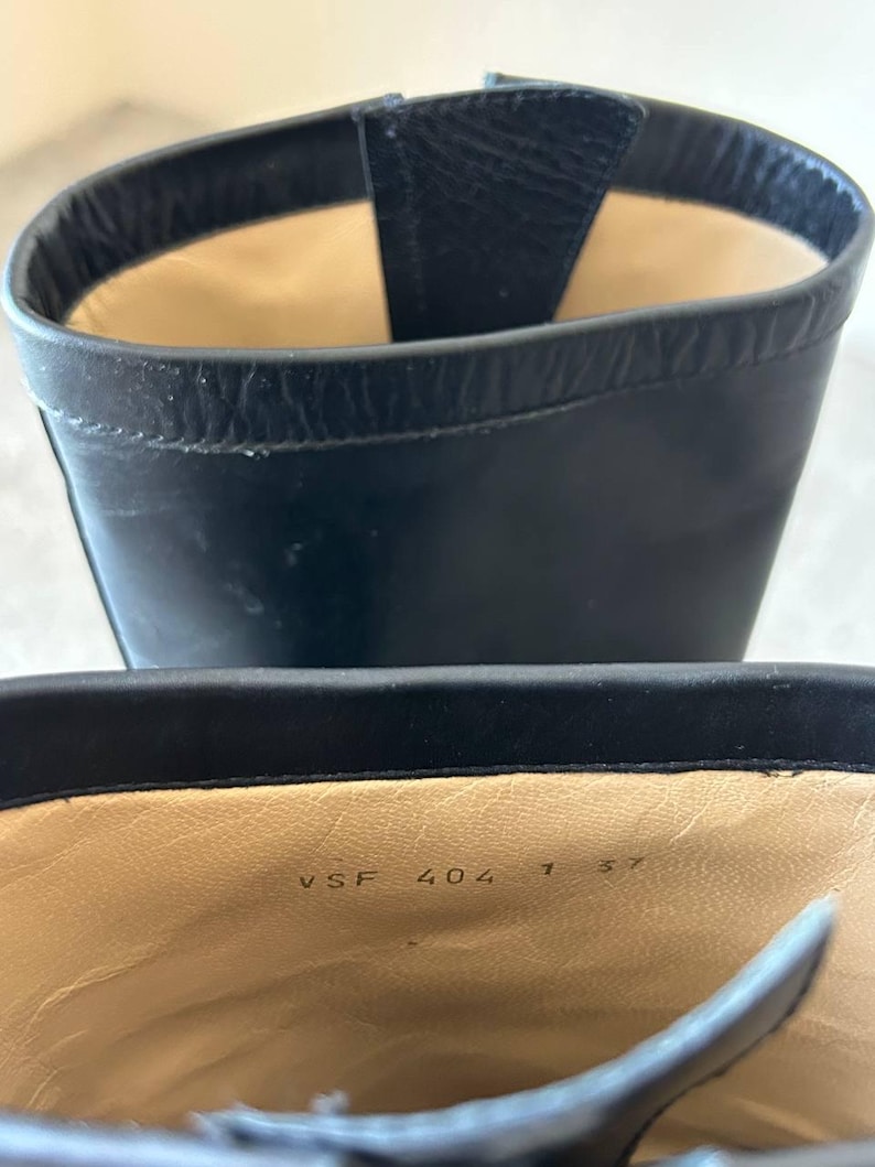 Peut inclure: Gros plan sur une botte en cuir noir. L'int&eacute;rieur est beige avec les chiffres "VSF 404 1 37" imprim&eacute;s sur la semelle int&eacute;rieure. L'ouverture de la botte est visible, r&eacute;v&eacute;lant la doublure int&eacute;rieure et les coutures. La botte est en cuir lisse et brillant.