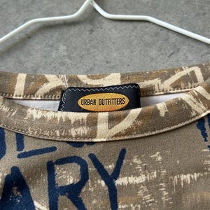 May include: Close-up of a crew neck shirt with a brown and beige abstract pattern and navy blue text. The shirt has a tag that reads "URBAN OUTFITTERS". The shirt is hanging on a white hanger.