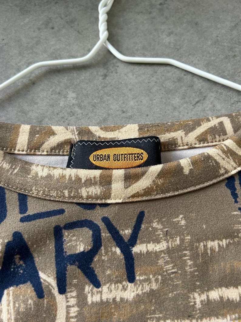May include: Close-up of a crew neck shirt with a brown and beige abstract pattern and navy blue text. The shirt has a tag that reads "URBAN OUTFITTERS". The shirt is hanging on a white hanger.