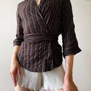 May include: Brown wrap-style blouse with vertical white pinstripes and a matching waist tie. The blouse has three-quarter sleeves and a wide collar. Paired with white pleated shorts.