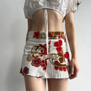 May include: A white crop top with short, puffed sleeves and a tie fastening. The mini skirt has a cream background and a bold floral pattern in red, brown, and gold. The skirt features a front slit.