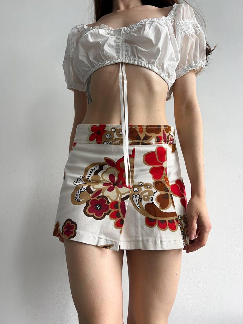 May include: A white crop top with short, puffed sleeves and a tie fastening. The mini skirt has a cream background and a bold floral pattern in red, brown, and gold. The skirt features a front slit.