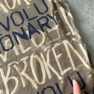 May include: Close-up of a fabric with a repeating word pattern. The words are in a mix of white and navy blue, with a brown background. The words include "BROKEN" and "REVOLUTIONARY".