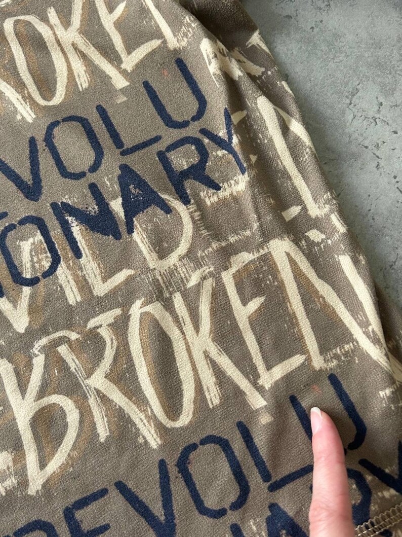 May include: Close-up of a fabric with a repeating word pattern. The words are in a mix of white and navy blue, with a brown background. The words include "BROKEN" and "REVOLUTIONARY".