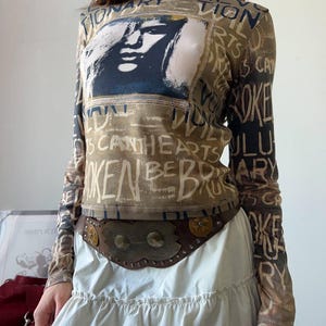 May include: A long-sleeved top with a graphic of a face and text. The top is brown with a white and black design. A brown belt with metal accents is worn over a white skirt.