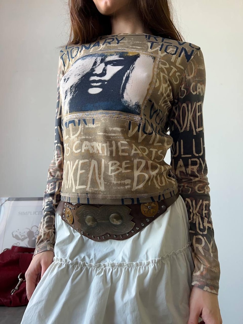 May include: A long-sleeved top with a graphic of a face and text. The top is brown with a white and black design. A brown belt with metal accents is worn over a white skirt.