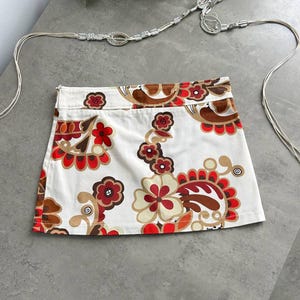 May include: A white mini skirt featuring a vibrant floral design in red, brown, and cream. The skirt has a straight cut and a visible zip. A silver belt with a decorative clasp is also visible.