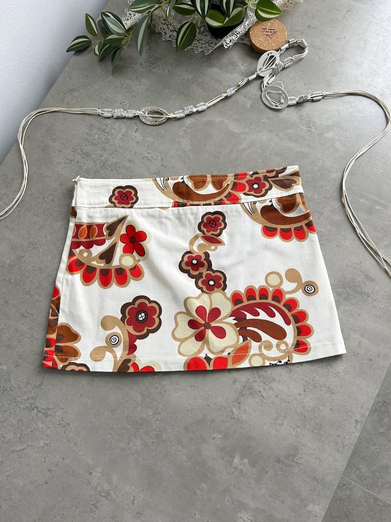 May include: A white mini skirt featuring a vibrant floral design in red, brown, and cream. The skirt has a straight cut and a visible zip. A silver belt with a decorative clasp is also visible.