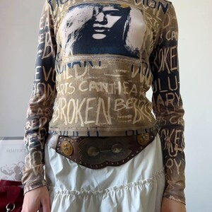 May include: A long-sleeved top with a graphic of a face and text in a distressed style. The top is brown and tan with black text. The text includes the words "BROKEN" and "REVOLUTIONARY". A brown leather belt with metal accents is worn over a white skirt.