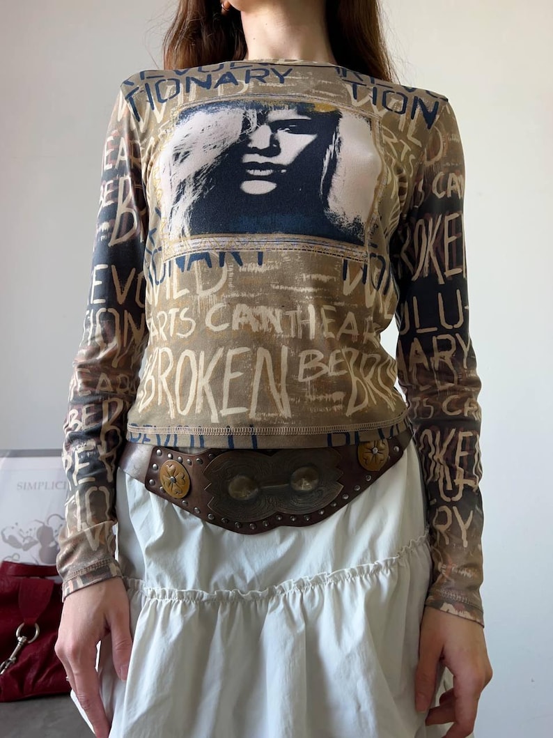 May include: A long-sleeved top with a graphic of a face and text in a distressed style. The top is brown and tan with black text. The text includes the words "BROKEN" and "REVOLUTIONARY". A brown leather belt with metal accents is worn over a white skirt.