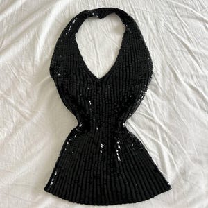 Op de afbeelding: Een zwarte haltertop met pailletten en een V-hals. De top is bedekt met verticale rijen zwarte pailletten, wat een gestructureerd, glinsterend effect creëert. Het kledingstuk wordt op een witte ondergrond getoond.