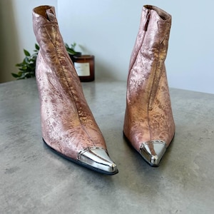 May include: Rose gold ankle boots with a metallic finish and silver pointed toes. The boots feature a high heel and a side zip. The textured material gives a unique look to the footwear.