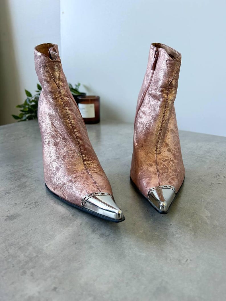 May include: Rose gold ankle boots with a metallic finish and silver pointed toes. The boots feature a high heel and a side zip. The textured material gives a unique look to the footwear.