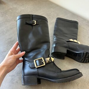 Peut inclure: Paire de bottes en cuir noir avec boucles dor&eacute;es et clous d&eacute;coratifs. Les bottes sont mi-mollet et ont un petit talon. Les bottes sont pr&eacute;sent&eacute;es sur une surface grise.