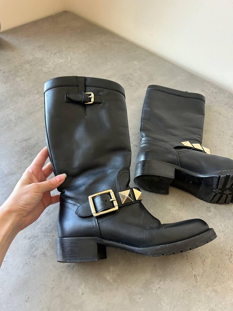 Peut inclure: Paire de bottes en cuir noir avec boucles dor&eacute;es et clous d&eacute;coratifs. Les bottes sont mi-mollet et ont un petit talon. Les bottes sont pr&eacute;sent&eacute;es sur une surface grise.