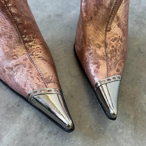 May include: Rose gold ankle boots with pointed toes and silver metal detailing. The boots have a metallic finish and a modern design. The silver metal detail is on the toe.