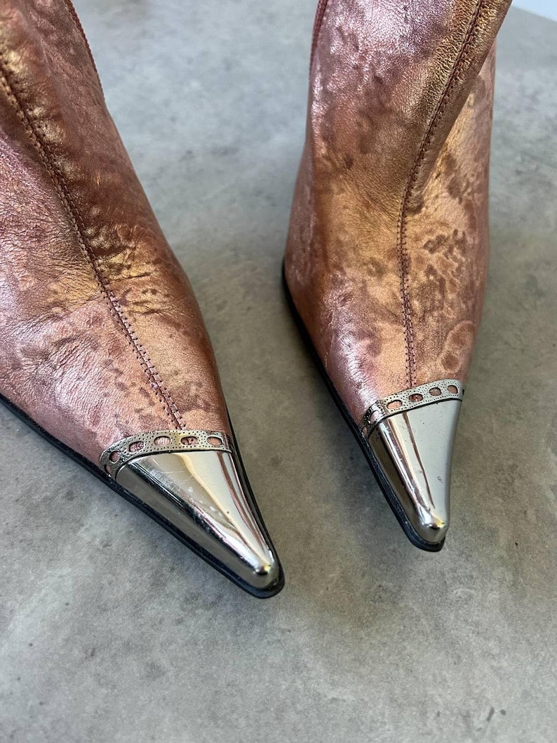 May include: Rose gold ankle boots with pointed toes and silver metal detailing. The boots have a metallic finish and a modern design. The silver metal detail is on the toe.