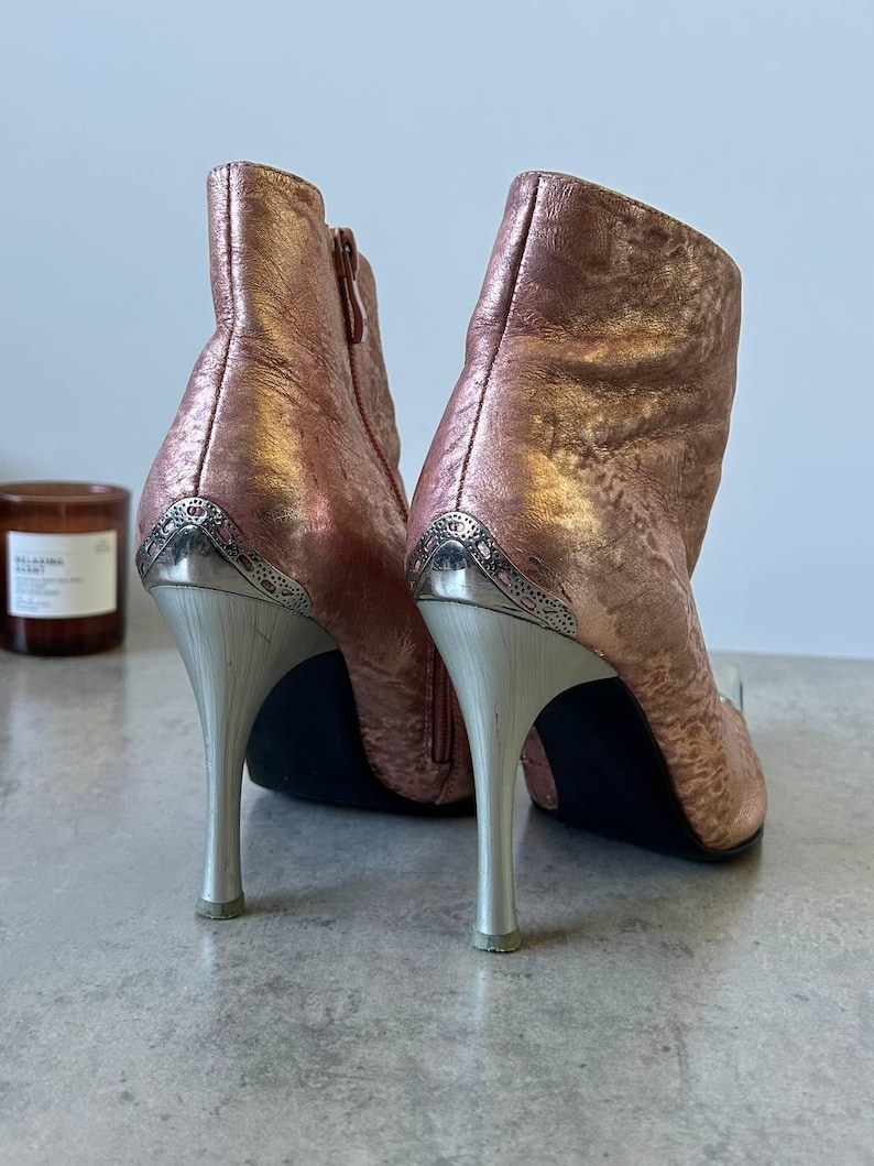 May include: Metallic rose-gold ankle boots with silver stiletto heels. The boots feature a decorative silver panel at the heel and a side zip. The heels are approximately 10 cm tall.