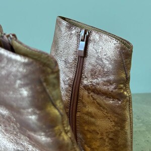 May include: Close-up of a metallic rose gold ankle boot. The boot features a side zip with a YKK zip pull. The leather has a textured, crinkled appearance. The boot is set against a light teal background.