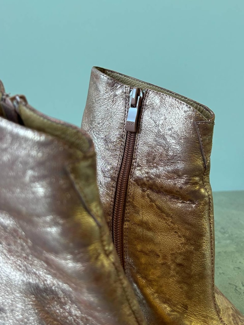 May include: Close-up of a metallic rose gold ankle boot. The boot features a side zip with a YKK zip pull. The leather has a textured, crinkled appearance. The boot is set against a light teal background.