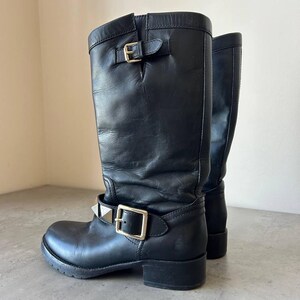 Peut inclure: Bottes hautes en cuir noir avec boucles argent&eacute;es et d&eacute;tails clout&eacute;s. Les bottes ont un bout rond et un petit talon. La tige des bottes est l&eacute;g&egrave;rement tombante. Un choix de chaussures &eacute;l&eacute;gant et audacieux.