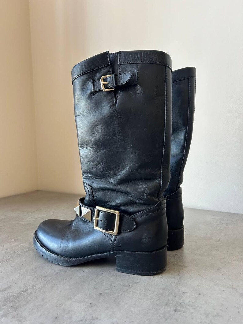 Peut inclure: Bottes hautes en cuir noir avec boucles argent&eacute;es et d&eacute;tails clout&eacute;s. Les bottes ont un bout rond et un petit talon. La tige des bottes est l&eacute;g&egrave;rement tombante. Un choix de chaussures &eacute;l&eacute;gant et audacieux.
