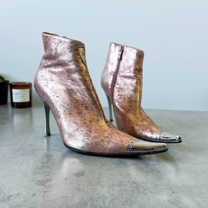 May include: Metallic rose-gold ankle boots with pointed toes and stiletto heels. The boots feature silver-coloured metal accents on the toes and zips on the inner sides. The heels are approximately 10 cm tall.