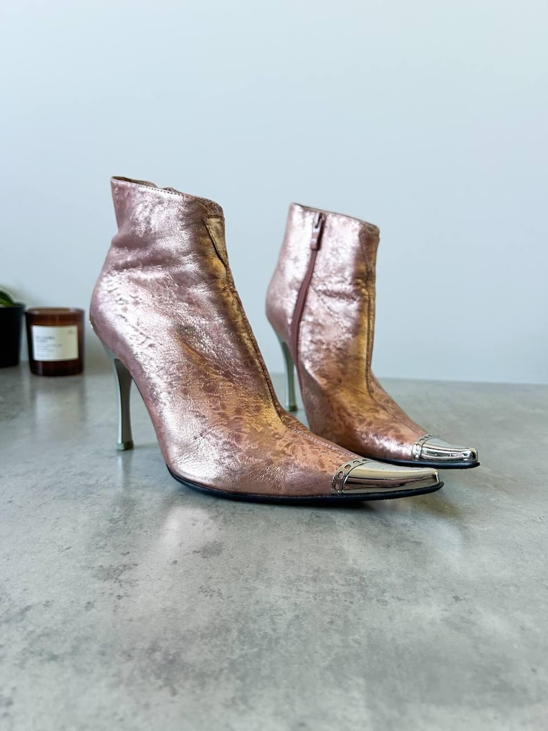 May include: Metallic rose-gold ankle boots with pointed toes and stiletto heels. The boots feature silver-coloured metal accents on the toes and zips on the inner sides. The heels are approximately 10 cm tall.