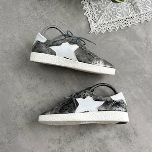 May include: A pair of metallic silver sneakers with a white star design on the side. The shoes have white soles and gray laces. The sneakers are on a gray surface.